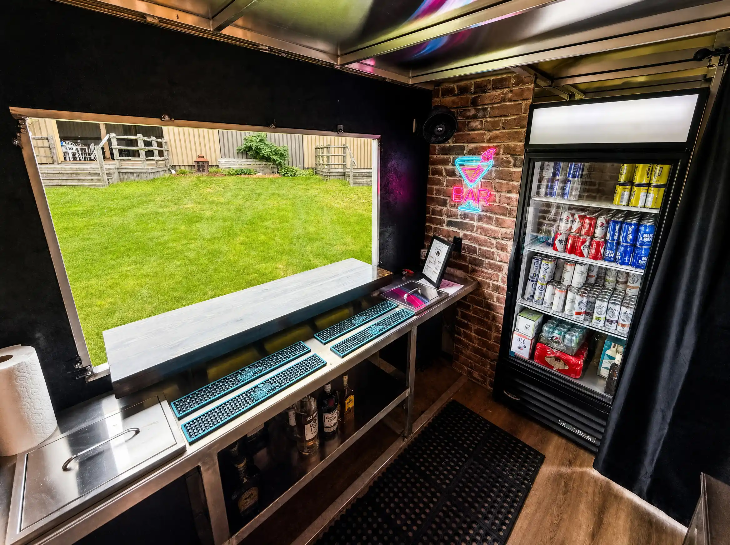 Inside the mobile bar, a glowing neon BAR sign above stocked fridges and the service window.
